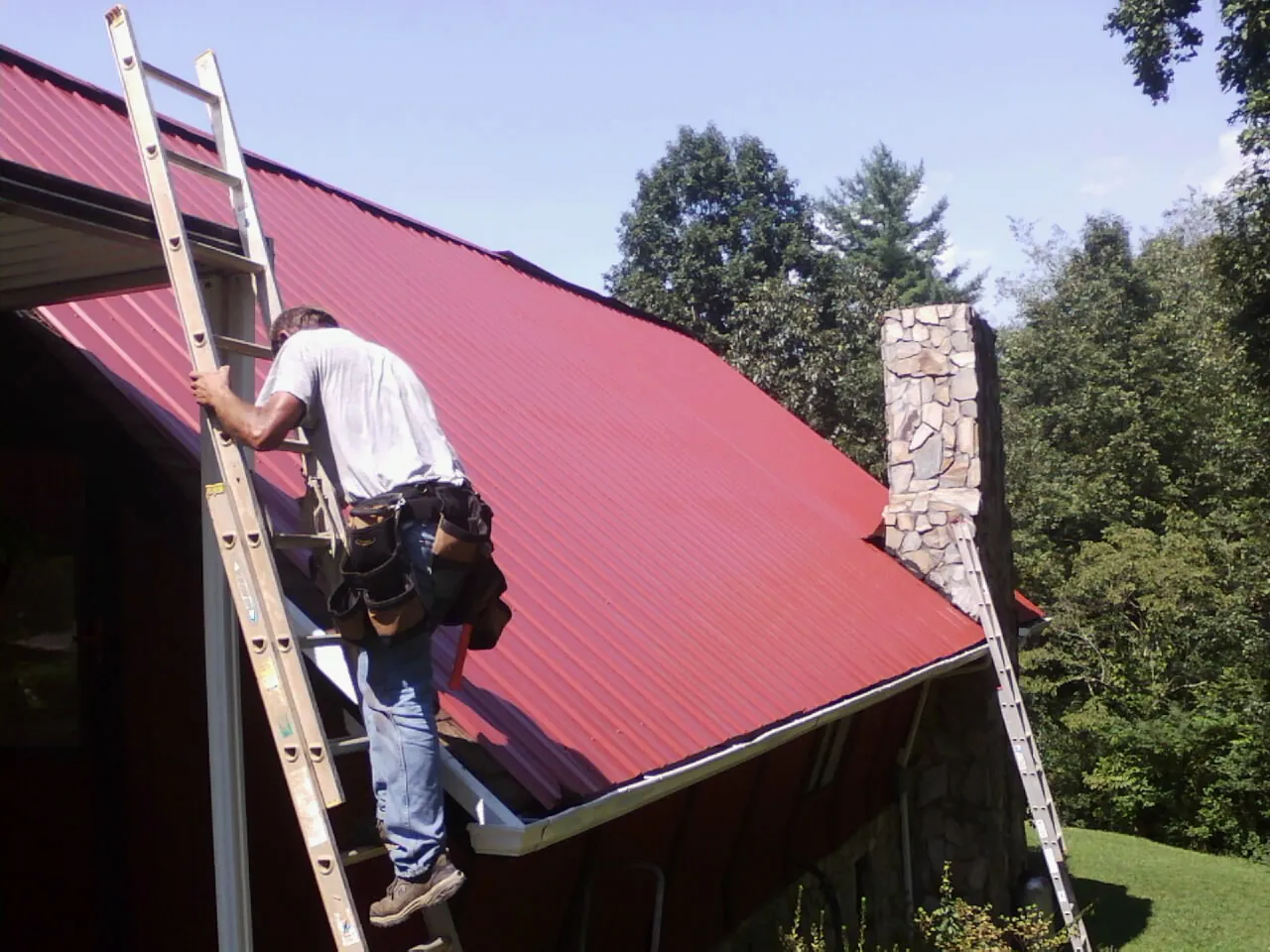Roofer on a ladder beside a red metal roof and stone chimney — Soffit & Fascia Repair by Roofing Brothers in Inglewood